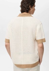 Polo din tricot cu mânecă scurtă, de culoare crem deschis, cu un model texturat și deschis, și guler și mansete ribbate de culoare bej.