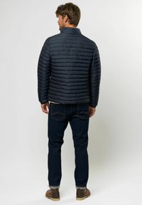 Marineblå pufferjakke med horisontalt quiltet stoff, høy krage og glidelås. Brukt over mørke jeans og brune støvler, og viser en avslappet passform.