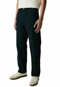 Tamsiai mėlynų džinsų kelnės su tiesiais koja, penkiomis kišenėmis ir užsegimu mygtuku, derinamos su kreminės spalvos sportbačiais.