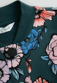 Ciemnozielony materiał z wielokolorowym kwiatowym nadrukiem, prążkowany kołnierz i duży czarny guzik z bliska.