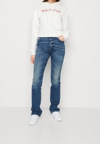 White hoodie with red graphic, paired with mid-rise blue jeans featuring a faded finish. White sneakers complete the outfit.