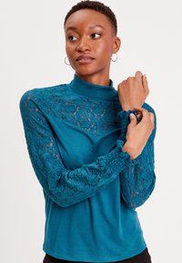 Maglietta a maniche lunghe color teal con colletto alto. Presenta maniche in pizzo con un motivo floreale e polsini arricciati. Realizzata in un morbido tessuto testurizzato.