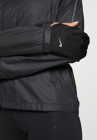Personne portant une veste de sport noire et un legging, la main partiellement couverte par un gant Nike noir montrant le logo.