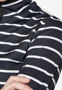 Primo piano di una persona che indossa una maglietta nera con strisce orizzontali bianche e un sottile logo "protect" sul petto.