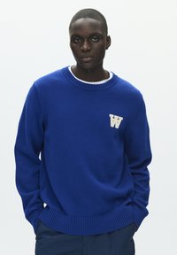 Pull en maille bleu avec poignets et ourlet côtelés, arborant un logo blanc "W" sur le côté gauche de la poitrine. Encolure ronde, texture douce.