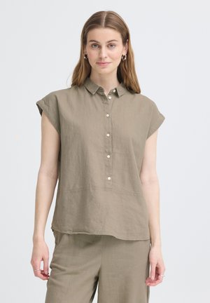 Kobieta ubrana w luźną, krótkawową, zapinaną na guziki taupe koszulę i dopasowane spodnie, stojąca na tle jasnego, jednolitego tła.