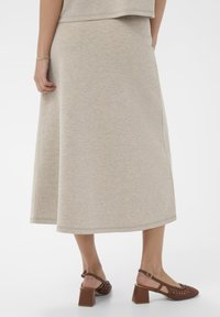 Bežs, līdz ceļiem garš svārki, kas izgatavoti no mīkstas, teksturētas auduma ar izplūdušu dizainu. Apvienoti ar brūnām blokveida papēžiem kurpēm ar austu akcentu.