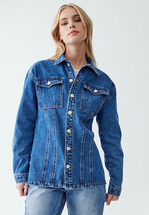 Chaqueta de mezclilla en azul medio, con mangas largas, cierre de botones en la parte delantera, dos bolsillos en el pecho y detalles de costura prominentes.