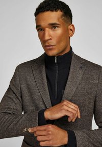 Brown tweed blazer with a textured finish, single chest pocket, and three-button cuff detail. Layered over a navy zip-up sweater.