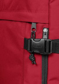 Röd ryggsäck med detaljer i texturerad tyg, med svarta dragkedjor, en clips-spänne och en dragsko med en knut.