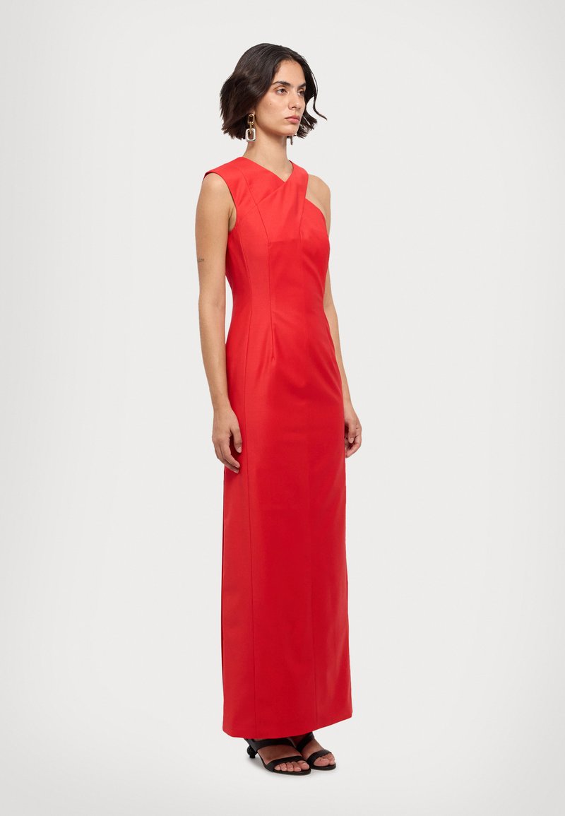 Robe maxi rouge sans manches avec un décolleté asymétrique et un corsage ajusté. Tissu lisse avec des coutures subtiles, associée à des sandales noires.