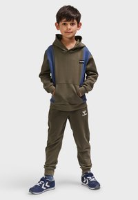 Olivgrüner Hoodie mit marineblauen Akzenten, Kängurutasche und Logo. Passende olivgrüne Jogginghosen mit elastischen Bündchen und dezentem Logodetail. Blaue Sneaker.