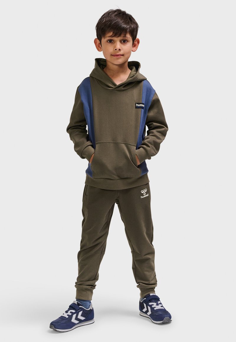 Olivgrüner Hoodie mit marineblauen Akzenten, Kängurutasche und Logo. Passende olivgrüne Jogginghosen mit elastischen Bündchen und dezentem Logodetail. Blaue Sneaker.