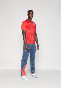 Rood sportshirt met een gestructureerd patroon en logo, gecombineerd met blauwe trainingsbroek met een roze accent. Witte en blauwe sportschoenen.