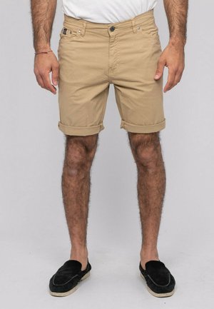 Homme portant un short beige retroussé, une chemise blanche et des chaussures noires sans lacets, debout devant un fond uni gris clair.