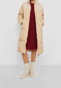 Beige pufjakke med en kremfarvet yoke, der har hætte og frontlynlås. Bæret over en burgunder strikkjole og parret med beige ankelstøvler.