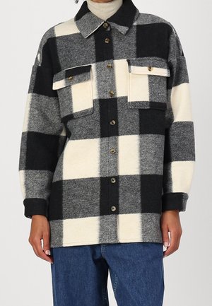 Person trägt eine schwarz-weiß-grau karierte Jacke mit Knöpfen, zwei Brusttaschen und blauen Jeans.