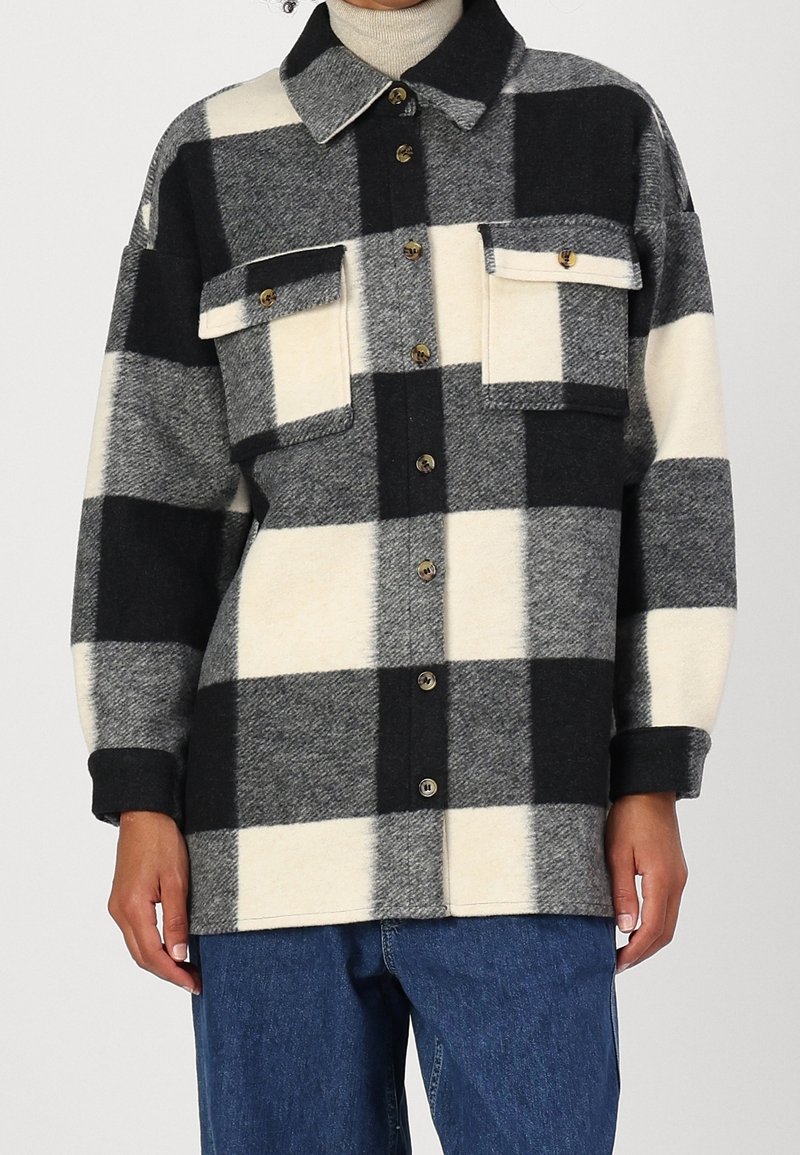 Personne portant une veste à grands carreaux noire, blanche et grise avec boutons, deux poches poitrine et un jean bleu.