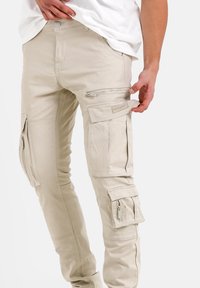Khaki spodnie cargo z wieloma zamykanymi na zamek i klapkami kieszeniami, wykonane z lekkiego materiału. Charakteryzują się dopasowanym krojem i widocznymi detalami szewnymi.