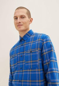 Niebieska koszula flanelowa w kratę z dwiema kieszeniami na piersi, kontrastującymi brązowymi i białymi paskami oraz kołnierzykiem zapinanym na guziki.