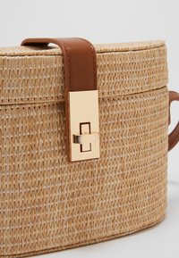 Borsa di paglia intrecciata rotonda di colore beige chiaro, con manico in pelle marrone e chiusura a scatto in metallo dorato per una chiusura sicura.