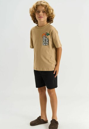 Beige cotton t-shirt with "ROAD TRIP" graphic in black, orange, and green; paired with black shorts and brown slip-on shoes.