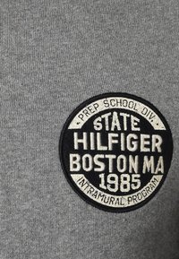 Grauer Baumwollmischstoff mit einem runden gestickten Patch. Der Patch enthält "STATE HILFIGER BOSTON MA 1985" und "INTRAMURAL PROGRAM" in kontrastierenden Farben.