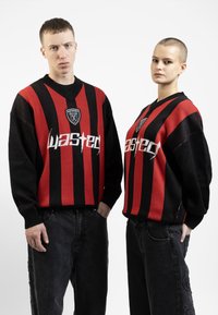 Sweaters surdimensionnés à rayures noires et rouges avec un col en V, présentant un logo "wasted" blanc bien visible et une patch sur le haut de la poitrine.