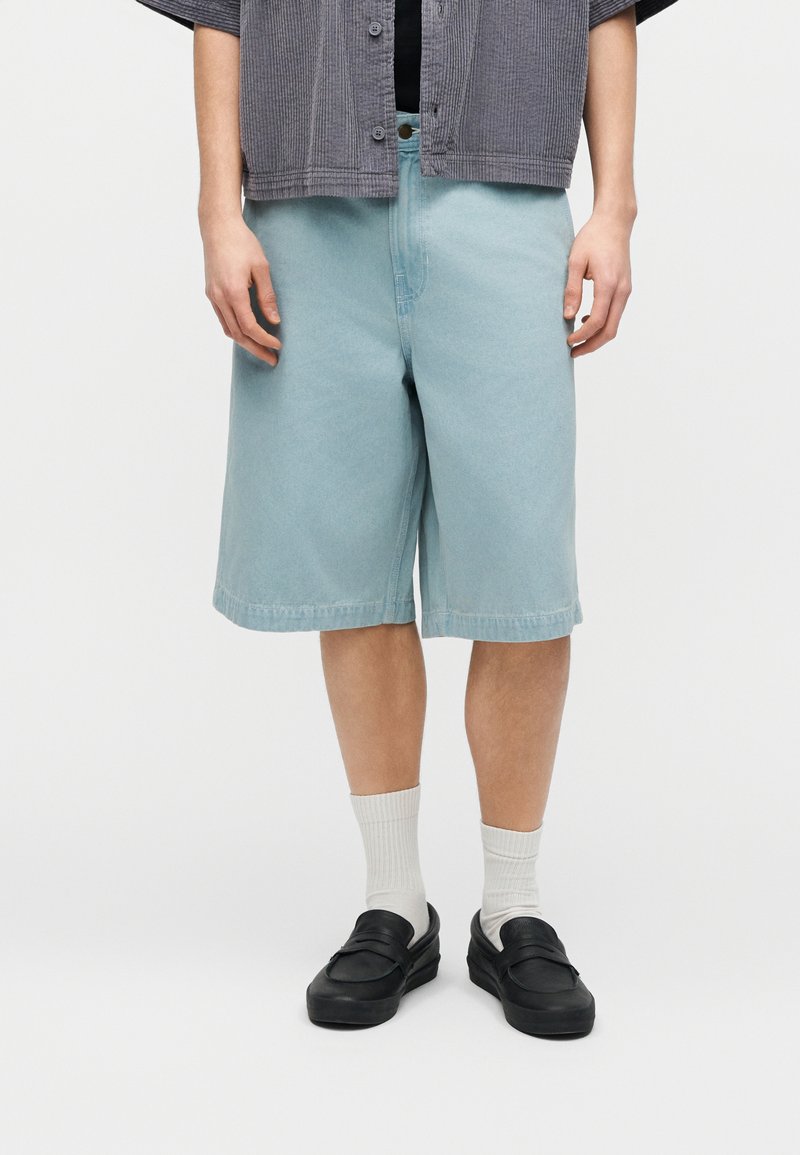 Person iført lyseblå denimshorts, hvide crew-sokker, sorte slip-on sko og en grå skjorte med knapper over en sort top.