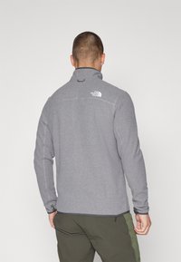 Grå fleecejakke med høj krave, sømdetaljer og et logo på ryggen. Designet inkluderer en trykknaplukning ved håndleddene.