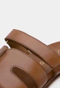 Καφέ δερμάτινο σανδάλι με δύο φαρδιές λωρίδες, λεία υφή και ορατές ραφές. Διαθέτει στρογγυλή μύτη και χρυσές λεπτομέρειες.