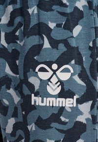 Blå kamouflage-mönstrad tyg med en texturerad design; vit logotyp "hummel" och emblem i mitten.