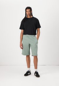 Μαύρο βαμβακερό t-shirt, κοντά μανίκια. Ανοιχτό πράσινο αθλητικό σορτς, ελαστική μέση, πλαινές τσέπες. Μαύρα αθλητικά παπούτσια με λευκές ρίγες, κάλτσες μέχρι τον αστράγαλο.