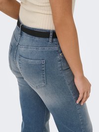 Persoon die lichtblauwe denim jeans en een beige gebreide mouwloze top draagt, weergegeven van de taille tot halverwege de dij, zijkant en achterkant te zien.