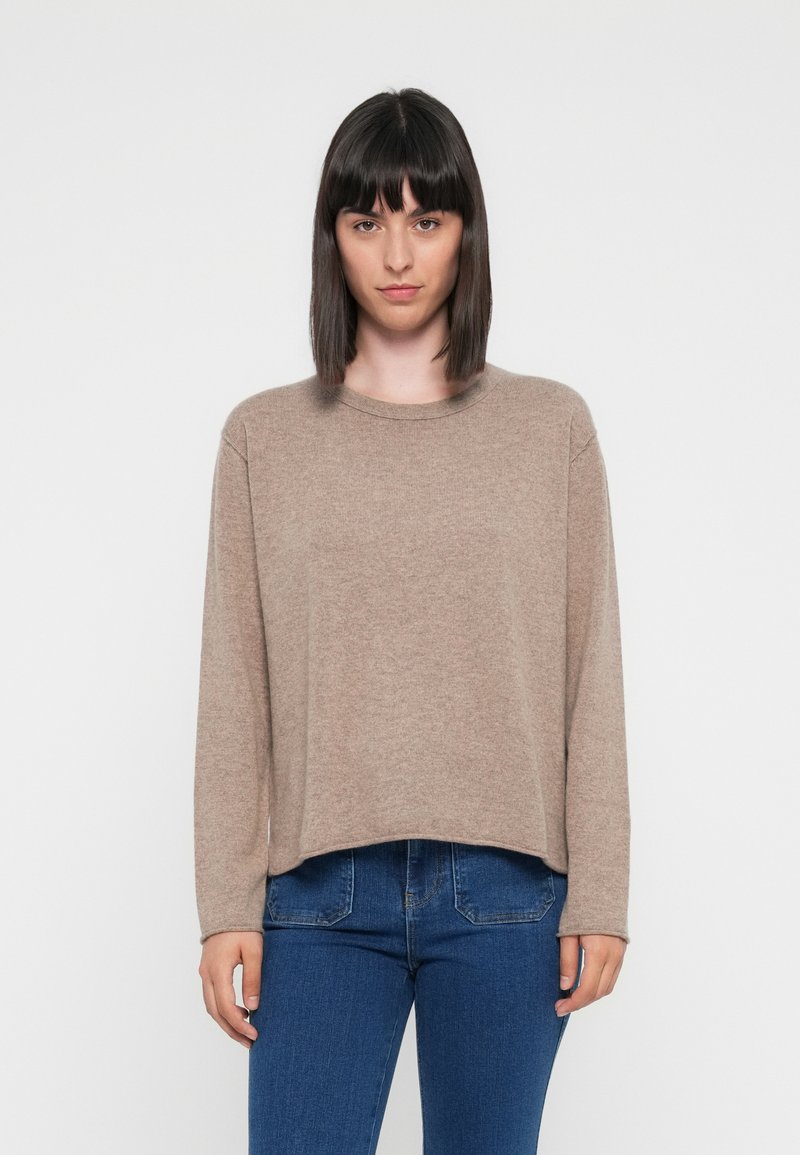 Beżowy sweter z długim rękawem, okrągłym dekoltem, teksturowanym materiałem i luźnym krojem, w zestawieniu z niebieskimi dżinsami. Prosty i casualowy design.
