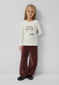 Langärmliges Creme-T-Shirt mit einer Ballerina-Grafik und dem Text "du kannst mich Prinzessin nennen", kombiniert mit weiten burgunderfarbenen Cordhosen und schwarzen Schuhen.