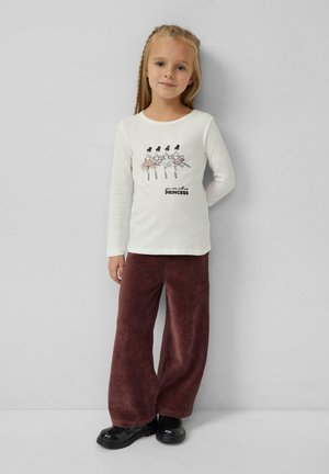 Langärmliges Creme-T-Shirt mit einer Ballerina-Grafik und dem Text "du kannst mich Prinzessin nennen", kombiniert mit weiten burgunderfarbenen Cordhosen und schwarzen Schuhen.