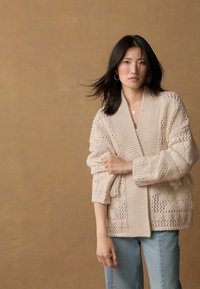 Světle béžový pletený cardigan s otevřeným předním dílem, texturovanými vzory a širokými rukávy, spojený se světle modrými džíny na neutrálním pozadí.