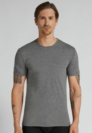 Mann mit hellbraunen Haaren, der ein schlichtes graues kurzärmeliges T-Shirt trägt, steht vor einem hellgrauen Hintergrund.