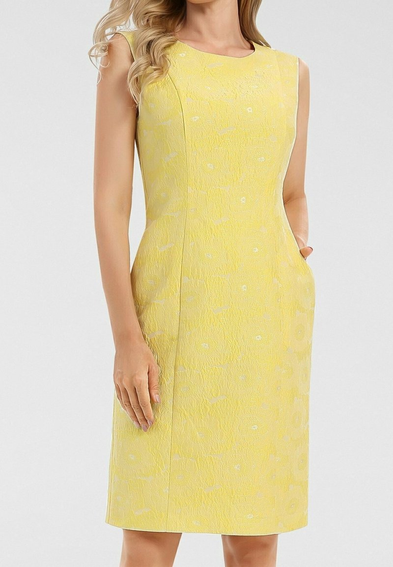 Robe jaune texturée sans manches, longueur genou, avec un motif floral subtil, portée par une femme aux cheveux blonds ondulés.