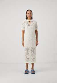 Robe midi en dentelle ivoire avec des manches courtes, un col à nouer, des motifs floraux et un ourlet à franges ; associée à des sandales slides bleues.