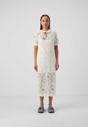 Robe midi en dentelle ivoire avec des manches courtes, un col à nouer, des motifs floraux et un ourlet à franges ; associée à des sandales slides bleues.
