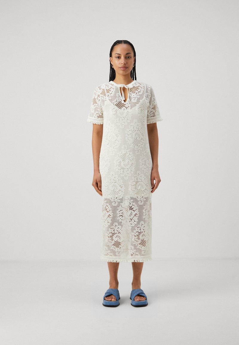Robe midi en dentelle ivoire avec des manches courtes, un col à nouer, des motifs floraux et un ourlet à franges ; associée à des sandales slides bleues.