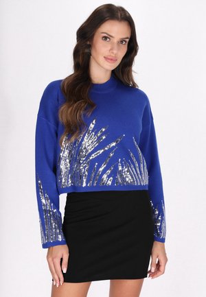 Frau mit langen braunen Haaren, die einen blauen Pullover mit silbernen Paillettenmustern und einen schwarzen Rock trägt, vor einem schlichten Hintergrund stehend.