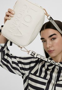 Giovane donna con una maglietta a righe bianche e nere tiene una borsa a tracolla bianca con impresso "LOLA CSDMNT" e un cinturino con il marchio.
