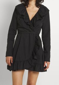 Vestido negro con mangas largas, escote en V profundo y acentos de volantes a lo largo del escote y el dobladillo. Diseño envolvente con una cintura ceñida y falda acampanada.