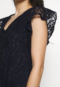 Marinblå spetsklänning med V-ringning och svävande ärmar. Tyget har blommönster och en mjuk, texturerad finish.