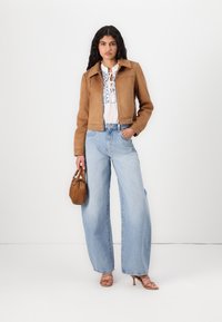 Brun fløyelslær kort jakke med nagledetaljer, hvit brodert bluse, høydejusterte lyseblå vide jeans, og en liten brun håndveske.