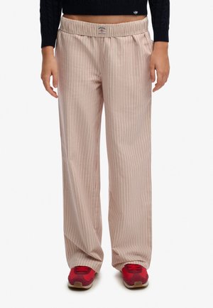 Kremowe spodnie z cienkimi czerwonymi pionowymi paskami, noszone z czerwonymi butami sportowymi i czarnym krótkim swetrem na stojącej osobie.