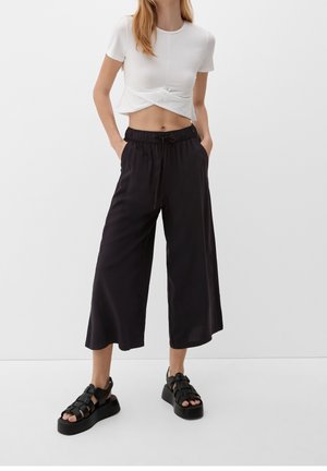 Kvinde iført hvid kortærmet crop-top, løst sorte vidde-ben kortbukser og sorte plateausandaler, stående med hænderne i lommerne.